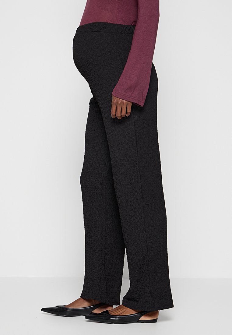Pantalones de maternidad negros texturizados con un corte relajado, que cuentan con una cintura elástica y piernas anchas. Combinados con una blusa de manga larga color marrón oscuro.