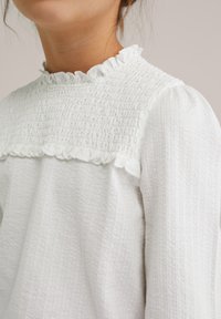 Blusa bianca con corpino smockato testurizzato e colletto arricciato. Presenta maniche lunghe con dettagli arricciati. Tessuto morbido e leggero.