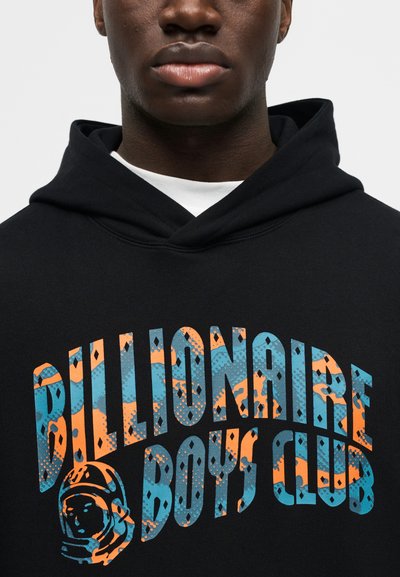 Κοντινό πλάνο ενός ατόμου που φοράει μαύρο φούτερ με κουκούλα, με το μπλε και πορτοκαλί λογότυπο "Billionaire Boys Club" και γραφικό ενός κράνους αστροναύτη στο στήθος.