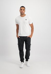 Man som står med händerna i fickorna, iklädd en vit kortärmad skjorta, svarta cargobyxor med lappar och vita sneakers mot en enfärgad bakgrund.