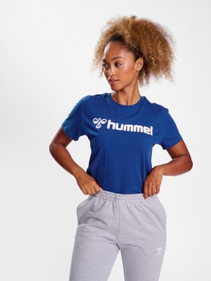 Kvinde med krøllet hår iført en blå Hummel t-shirt og lysegrå joggingbukser, justerer taljen, på en ensfarvet hvid baggrund.