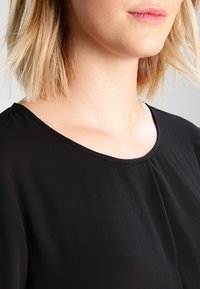 Blusa nera con scollatura tonda e leggere pliche. Texture morbida e liscia, con maniche corte. Inquadratura focalizzata sulla parte superiore del corpo, senza dettagli aggiuntivi.