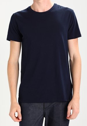 Person iført en ensfarvet mørkeblå kortærmet t-shirt og mørkegrå bukser, stående mod en lys baggrund.