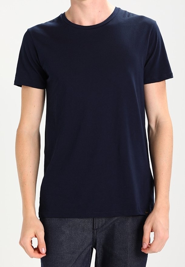 Personne portant un t-shirt uni bleu marine à manches courtes et un pantalon gris foncé, se tenant devant un fond clair.