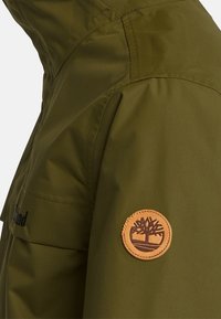 Oliivinvihreä takki, jossa on vedenpitävä materiaali, saumaus yksityiskohtia ja pyöreä, vaaleanruskea nahkalogo-patch hihassa.