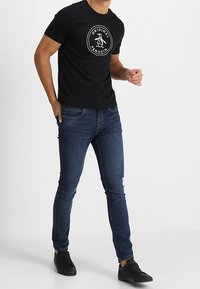 Hombre vistiendo una camiseta negra de "Original Penguin", jeans ajustados azul oscuro y zapatillas negras, de pie frente a un fondo blanco liso.