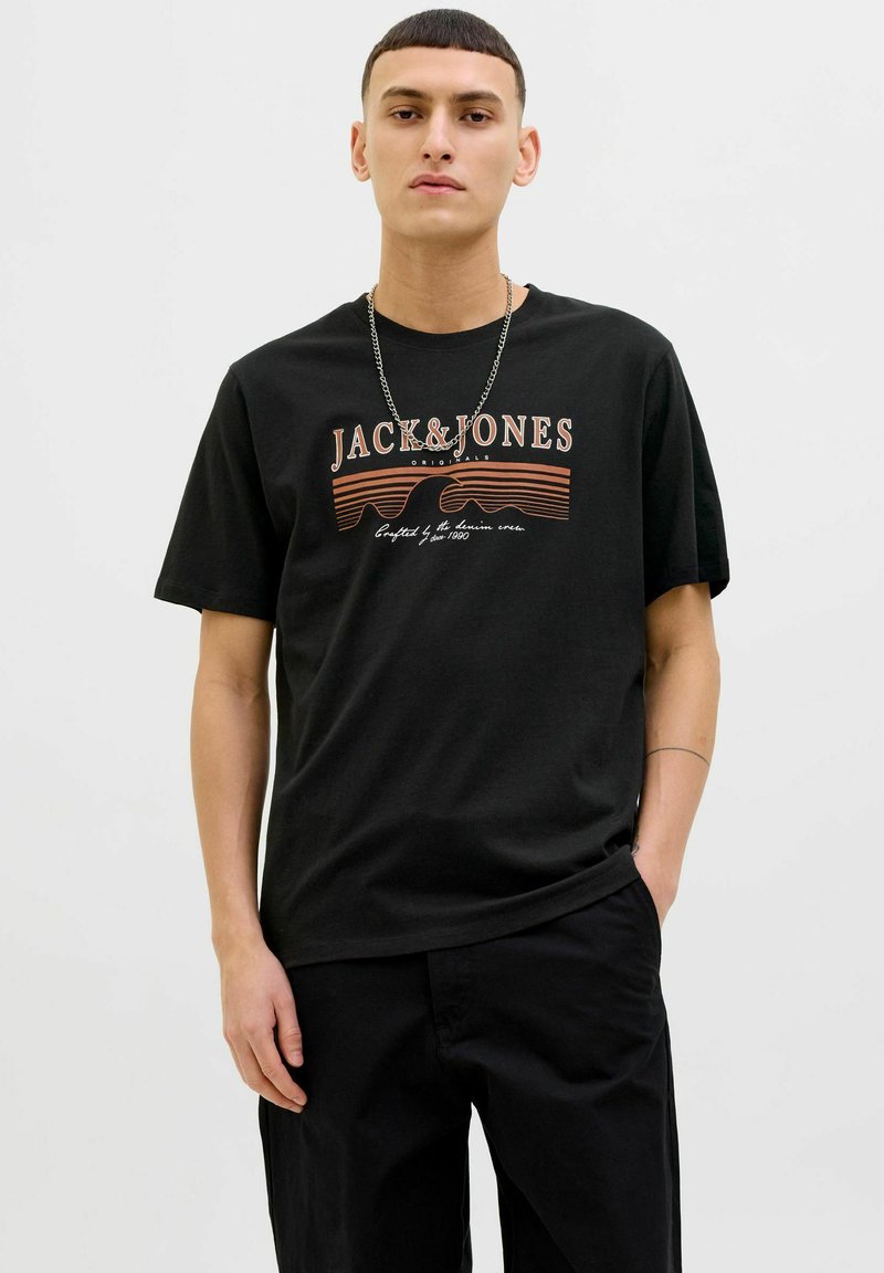 Melna kokvilnas T-kreklu ar priekšējo grafiku, kurā redzams "JACK & JONES" baltā un oranžā tekstā, apvienots ar brīva piegriezuma melnajām biksēm.