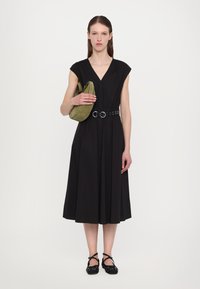 Abito nero senza maniche con scollatura a V, accentuato da una cintura larga, abbinato a una borsa a tracolla verde e scarpe nere con cinturini incrociati.