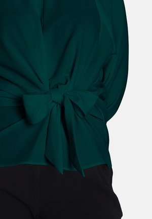Bluză teal cu un design plisat și detaliu de nod în spate. Materialul pare ușor, cu o textură netedă. Parte din fundalul negru este parțial vizibil.