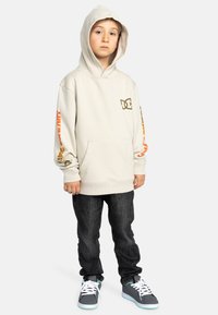 Ljusbeige hoodie med en framficka, med orange och svarta grafiska detaljer på ärmarna. Matchad med mörka jeans och grå sneakers.