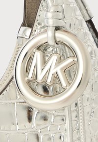 Detail stříbrné kabelky z krokodýlí kůže s embosovaným vzorem, která obsahuje kulatou kovovou visačku s logem "MK" a uzlovou rukojeť.