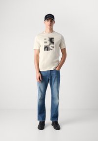 Man wearing a beige graphic T-shirt, blue jeans, black sneakers, and a black cap standing against a plain white background.