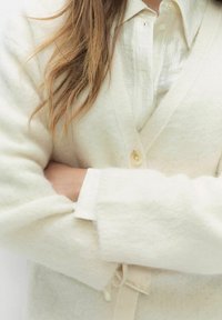 Kremowy kardigan wykonany z miękkiej, strukturalnej wełny, z dekoltem w kształcie litery V, z pojedynczym zapięciem na guzik, noszony na jasnej koszuli z kołnierzykiem.