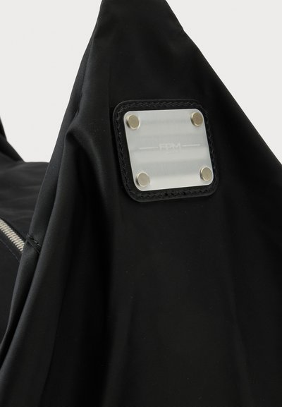 Μαύρο ύφασμα με ασημένια μεταλλική τετράγωνη πλάκα στερεωμένη με τέσσερις νίτες, προσαρτημένη σε μαύρο ραμμένο λεκέ σε μια τσάντα ή ένδυμα.