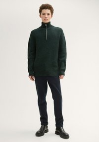 Donkergroen gebreide trui met hoge kraag en zilveren rits, gecombineerd met marineblauwe broek en zwarte chunky laarzen. Gladde textuur, geribbeld patroon.