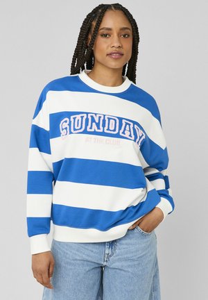 Femme aux cheveux tressés portant un sweat-shirt à rayures bleues et blanches avec le texte "SUNDAY AT THE CLUB" et un jean bleu clair, debout devant un fond gris.