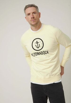 Hombre con sudadera amarillo pálido con logo de ancla negra y texto "ALTONADOCK", posando con las manos detrás de la espalda sobre un fondo liso.