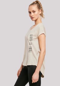 Beige Baumwoll-T-Shirt mit kurzen Ärmeln, rundem Halsausschnitt und einem leicht verlängerten Rücken. Mit vertikalem schwarzem Text "TAKE IT EASY."