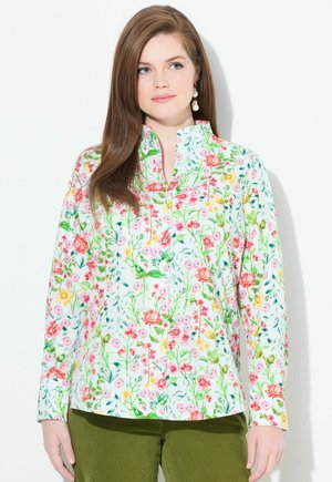 Mujer con cabello largo y castaño que lleva una camisa blanca de botones con un estampado floral colorido y pantalones verde oliva, de pie contra un fondo blanco.