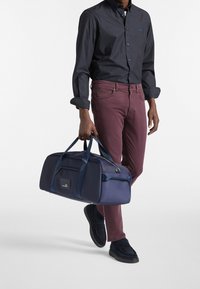 Marineblauwe reistas van duurzaam materiaal, met dubbele handgrepen en een logo-patch. Het model draagt een donker shirt met patroon en bordeauxrode broek, gecombineerd met donkere schoenen.