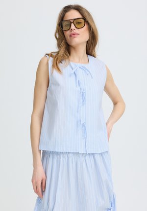 Femme portant un chemisier bleu clair rayé sans manches avec des nœuds et une jupe assortie, posant la main sur la hanche et portant des lunettes teintées marron.