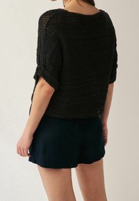 Czarny sweterek z krótkim rękawem, charakteryzujący się luźnym krojem i teksturowanym wzorem. Noszony z ciemnymi szortami, co podkreśla swobodny styl.