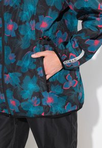Blommig jacka med svart bakgrund, har blå och röda blommönster, dragkedjefickor, justerbara ärmslut och en texturerad tyg.