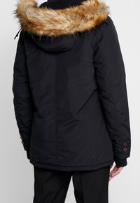 Svart parka-jacka med fuskpälsfodrad huva, med sidofickor och knappdetaljer vid ärmsluten. Slät, isolerad textil.