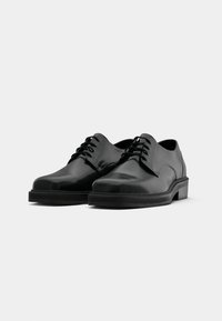 Chaussures en cuir noir à lacets avec un bout rond, texture lisse, semelle plate et design minimaliste. Pas de logos visibles ni d'accents décoratifs.