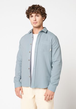 Jeune homme aux cheveux bruns bouclés portant une chemise à rayures bleues claires sur un t-shirt blanc et un pantalon crème, debout devant un fond blanc.