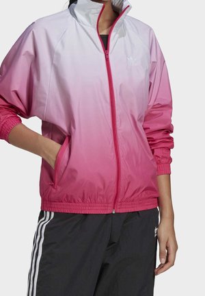 Person trägt eine Zip-Up-Jacke mit pink-weißem Farbverlauf und elastischen Bündchen sowie schwarze Hosen mit weißen Streifen.
