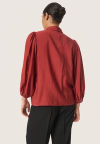 Red satin blouse with a relaxed fit, featuring puffed sleeves and a classic collar, paired with black trousers. Smooth texture, minimal detailing.