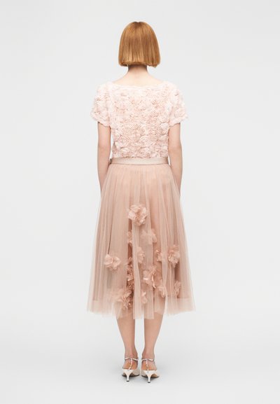 Γυναίκα με κοντά κόκκινα μαλλιά που φοράει ροζ ανοιχτό τοπ με άνθη και διάφανη ροζ φούστα στολισμένη με υφασμάτινα λουλούδια, γυρισμένη πίσω.