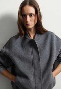 Veste en laine grise surdimensionnée avec un col haut, dotée d'une fermeture à bouton et de poignets côtelés. Design épuré et minimaliste, texture douce.