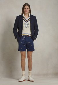 Jūras spēku saspringtā jakā virs baltas apkakles krekla, krēmkrāsas adīts džemperis ar ziliem svītrojumiem, tumši zilas šortas ar izšūtiem tenisa raketēm, baltas apavas.