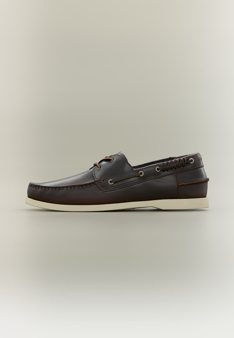 Chaussure bateau en cuir marron foncé avec semelle blanche, couture moc toe, œillets pour lacets et laçage décoratif sur le côté, sur un fond neutre.