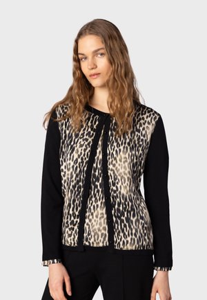 Kvinde med langt bølget hår iført en knaplukket leopardprint cardigan med sorte ærmer og sorte bukser mod en ensfarvet lys baggrund.
