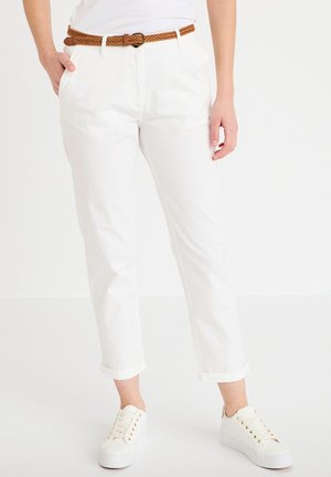 Person iført hvide korte bukser med opkørte kanter, et flettet brunt bælte og hvide platform-sneakers, stående mod en hvid baggrund.