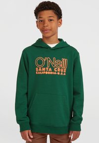 Gutt med kort krøllet hår iført en grønn O'Neill Santa Cruz hettegenser og brune bukser, stående mot en ensfarget grå bakgrunn.