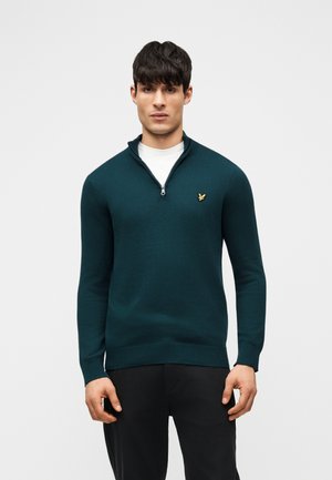 Fiatal férfi sötét tengerzöld negyed cipzáras pulóvert visel fehér aláöltözet és fekete nadrág felett, semleges világos háttér előtt.