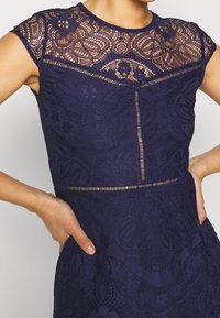 Krajkové šaty v tmavě námořnické modré barvě s detailním krajkovým živůtkem, krátkými rukávy a přiléhavým pasem s texturovaným vzorem a průsvitnými prvky.