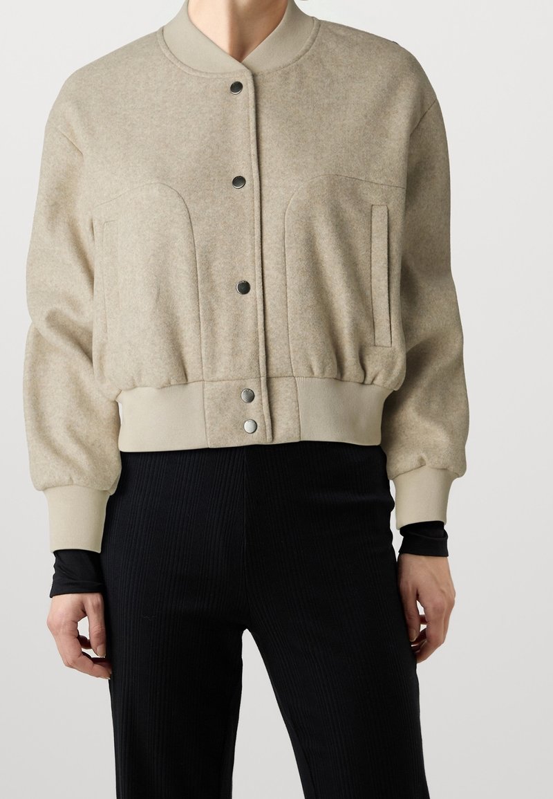 Beżowa bomberka z wełny z ryflowanym dołem, zapięciem na napy i dwoma bocznymi kieszeniami; ma teksturowane wykończenie oraz kontrastowy kołnierz.