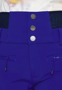 Paarse high-waisted broek met smalle riem en drie metalen knopen, voorzien van voorzakken met ritssluiting en een textuurdetail op de tailleband.