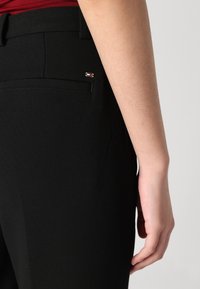 Black tailored trousers with a smooth texture, featuring a back pocket and a small logo accent. The waistband is flat and streamlined.