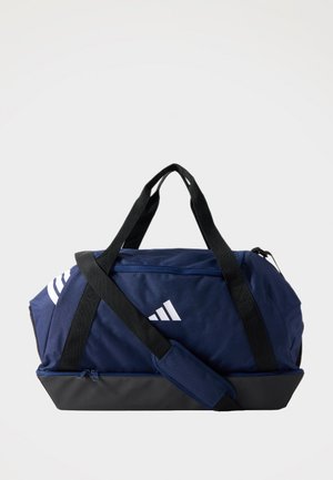 Tengerészkék és fekete utazótáska fekete fogantyúkkal és vállpánttal, fehér Adidas logóval és cipzáras rekeszekkel, egyszínű háttéren.