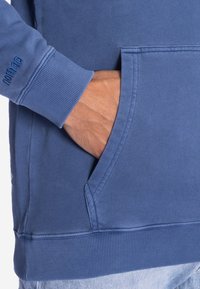 Blauer Kapuzenpullover mit Kängurutasche, gerippten Bündchen und Saum. Das Material wirkt weich, mit sichtbaren Nähten und einem Logo auf dem Ärmel.