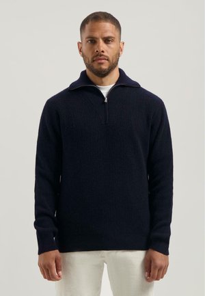 Marineblauwe gebreide pullover met een kwart ritssluiting, geribbelde textuur en een hoge kraag, gecombineerd met lichtgekleurde corduroy broeken.