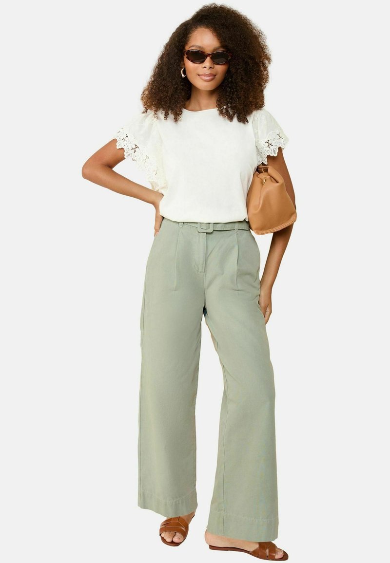 Blusa di crema con maniche in pizzo, pantaloni larghi verde chiaro, borsa marrone e sandali con cinturini marroni. La modella ha capelli ricci e occhiali da sole.