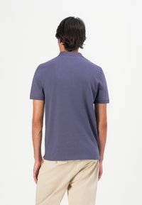 Chemise polo violette à manches courtes, avec un col classique et une texture douce. Associée à un pantalon beige, vue de dos.
