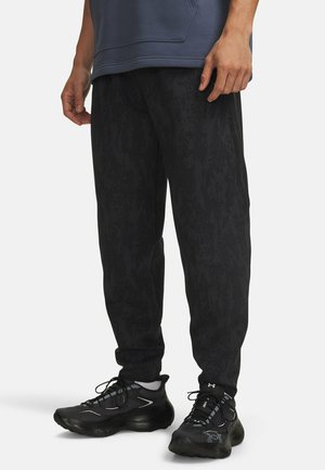 Man draagt zwarte joggingbroek met patroon en zwarte sportschoenen met witte sokken, staand tegen een effen achtergrond.